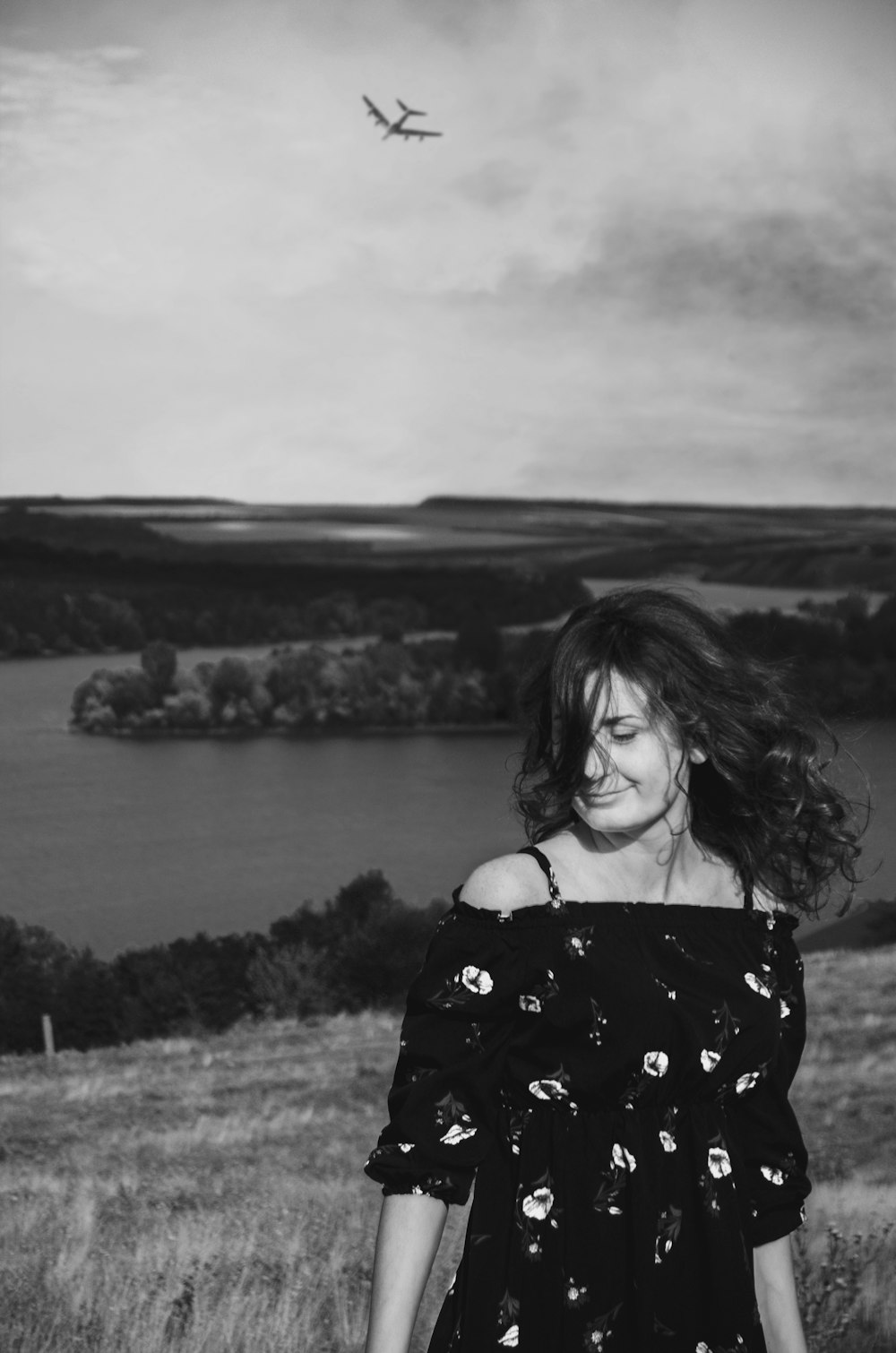 woman standing on hill top near lake with airplane flying above