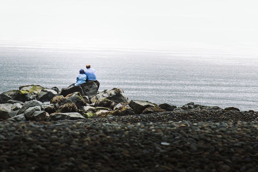 파란 셔츠를 입은 남자와 여자가 해변의 바위에 앉아 있다