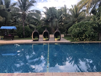 A serene swimming pool is surrounded by lush tropical trees. Three woven pod chairs with green cushions sit on the poolside deck. A cart with a blue canopy is situated on the grass near the chairs.