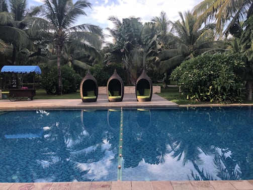 A serene swimming pool is surrounded by lush tropical trees. Three woven pod chairs with green cushions sit on the poolside deck. A cart with a blue canopy is situated on the grass near the chairs.