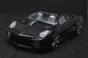 A sleek, angular, black sports car model with a shiny exterior and metallic accents on the wheels and body. The car is positioned against a dark backdrop, highlighting its glossy finish and aerodynamic design.