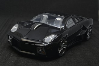 A sleek, angular, black sports car model with a shiny exterior and metallic accents on the wheels and body. The car is positioned against a dark backdrop, highlighting its glossy finish and aerodynamic design.