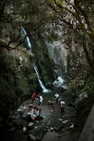 A group of people are gathered at the base of a small waterfall surrounded by lush greenery and large rocks. The water flows down the rocky cliff into a clear pool. Some individuals are standing in the water while others relax on the stones nearby. Sunlight filters through the branches above, creating a serene natural setting.