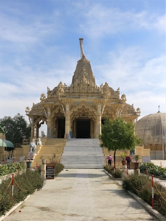 An ornately designed temple with intricate carvings and sculptures on its fa&ccedil;ade. The central entrance features a grand staircase leading up to the detailed main sanctuary. The temple is surrounded by greenery and additional architectural structures, with a clear blue sky overhead.