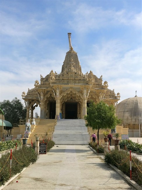 An ornately designed temple with intricate carvings and sculptures on its fa&ccedil;ade. The central entrance features a grand staircase leading up to the detailed main sanctuary. The temple is surrounded by greenery and additional architectural structures, with a clear blue sky overhead.