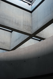 Concrete walls intersect in a geometric architectural design, with lines and shadows creating a modern aesthetic. The lighting from above highlights the smooth texture of the surfaces, emphasizing the structural form.