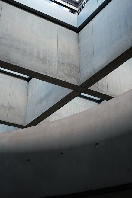 Concrete walls intersect in a geometric architectural design, with lines and shadows creating a modern aesthetic. The lighting from above highlights the smooth texture of the surfaces, emphasizing the structural form.