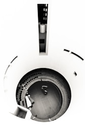 A circular structure with concentric steps leading down to a central area, featuring a small vehicle or machinery piece. There are scattered objects, including a covered item resembling furniture and a row of circular elements. The walls are smooth and curving, giving a minimalist and modern aesthetic.