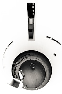 A circular structure with concentric steps leading down to a central area, featuring a small vehicle or machinery piece. There are scattered objects, including a covered item resembling furniture and a row of circular elements. The walls are smooth and curving, giving a minimalist and modern aesthetic.