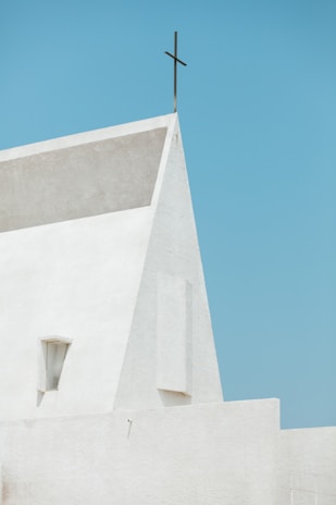 A minimalist architectural structure with a modern, triangular facade, topped by a simple cross against a clear blue sky.