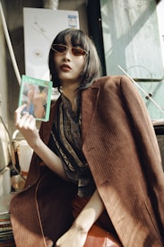 A person with short dark hair wearing dark sunglasses and a stylish brown coat sits inside a room. They hold a colorful magazine or book, which features a nude painting or photograph. The lighting creates soft shadows, and there is a hint of a vintage aesthetic in the decor.
