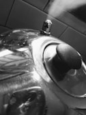 A shiny metal kettle with steam emitting from its spout is sitting on a stovetop. The black handle and lid are visible, and the tiled wall in the background provides contrast to the reflective surface of the kettle.