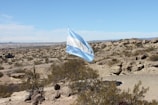 A flag featuring blue and white horizontal stripes is waving prominently in a vast, arid landscape. The terrain is rocky with sparse vegetation and cacti scattered throughout the scene. The sky is clear with a few clouds visible in the distance.