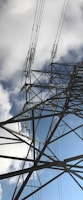 A towering metal electricity pylon extends upwards, with high tension wires stretching into the cloudy sky. Diagonal beams and insulators are visible, creating a geometric pattern against a backdrop of blue sky partially obscured by clouds.