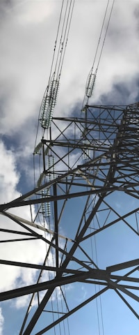 A towering metal electricity pylon extends upwards, with high tension wires stretching into the cloudy sky. Diagonal beams and insulators are visible, creating a geometric pattern against a backdrop of blue sky partially obscured by clouds.