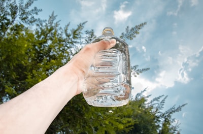 A hand is holding up a clear plastic container filled with water against a backdrop of a partly cloudy blue sky and green tree foliage.