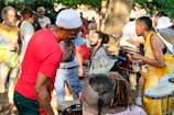 A lively outdoor gathering with people playing drums and percussion instruments. Participants are dressed in colorful attire and appear to be enjoying the music and atmosphere. The background shows a mix of trees and bystanders lightly engaging with the scene.