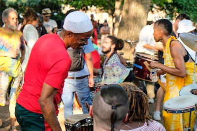 A lively outdoor gathering with people playing drums and percussion instruments. Participants are dressed in colorful attire and appear to be enjoying the music and atmosphere. The background shows a mix of trees and bystanders lightly engaging with the scene.