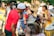 A lively outdoor gathering with people playing drums and percussion instruments. Participants are dressed in colorful attire and appear to be enjoying the music and atmosphere. The background shows a mix of trees and bystanders lightly engaging with the scene.