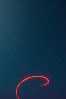 A night sky filled with numerous small stars scattered across a deep blue background. A bright, swirling red light trail stands out against the darkness, creating a captivating spiral shape near the bottom of the image.