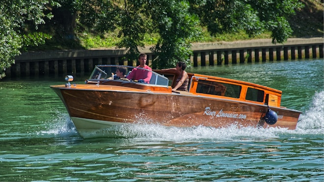 A wooden motorboat is gliding through a body of water, causing small waves and splashes. The boat is marked with 'River Limousine.com' on its side. Two people are seated at the front under a clear sky, enjoying the ride. The backdrop includes lush green trees and a wooden retaining wall lining the water's edge.