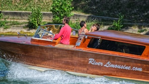 A wooden boat with the name 'River Limousine.com' is moving quickly on the water, creating waves. Three people are visible on board; one is steering the boat, and the other two are seated, engaged in conversation. The background shows a grassy and tree-lined riverside.