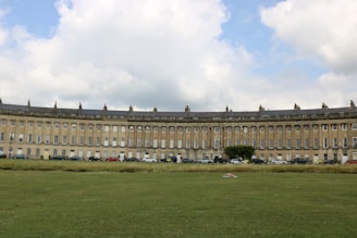 A large, curved row of Georgian terrace houses is seen against a backdrop of a partly cloudy sky. The architecture features tall columns and uniform windows, typical of historical design. In the foreground, a wide expanse of green lawn stretches out, with a few cars parked along the street in front of the buildings.