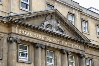 A historic stone building facade with classical architectural elements. An ornate pediment features a heraldic emblem and the inscription 'Royal Mineral Water Hospital'. Large, symmetrical windows are supported by decorative columns.