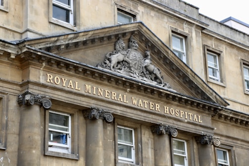 A historic stone building facade with classical architectural elements. An ornate pediment features a heraldic emblem and the inscription 'Royal Mineral Water Hospital'. Large, symmetrical windows are supported by decorative columns.