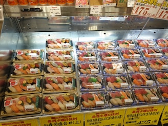 A supermarket display case features a variety of pre-packaged sushi trays. Each tray contains an assortment of sushi, with toppings like salmon, tuna, and shrimp carefully arranged. Japanese labels and price tags are visible on each tray, and the display is well-lit to enhance the freshness and appeal of the sushi offerings.