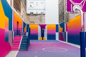 A vibrant, multicolored urban basketball court nestled between buildings. The walls and floor are decorated with bold geometric patterns in shades of pink, purple, blue, and yellow. The court features a basketball hoop and a set of stairs leading to a door.