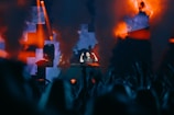 A DJ is performing on stage amidst a lively atmosphere, with vibrant lights and smoke effects creating a dynamic backdrop. The audience's silhouettes can be seen in the foreground, enhancing the energetic ambiance.