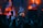 A DJ is performing on stage amidst a lively atmosphere, with vibrant lights and smoke effects creating a dynamic backdrop. The audience's silhouettes can be seen in the foreground, enhancing the energetic ambiance.