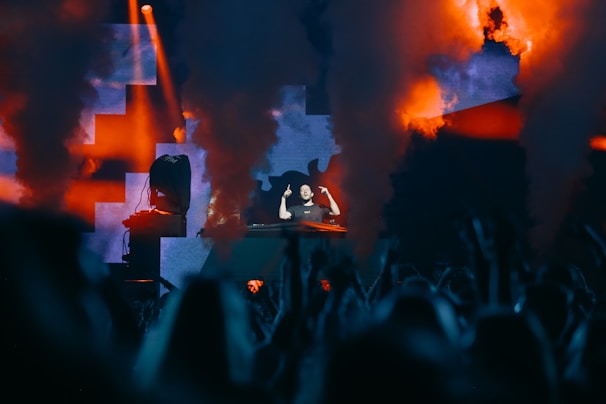 A DJ is performing on stage amidst a lively atmosphere, with vibrant lights and smoke effects creating a dynamic backdrop. The audience's silhouettes can be seen in the foreground, enhancing the energetic ambiance.
