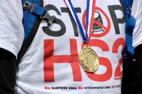 A person is wearing a white shirt with a bold message against HS2, featuring a red circle crossed out over a train. They have a gold medal hanging around their neck on a ribbon. A blue strap or bag is visible over their shoulder.