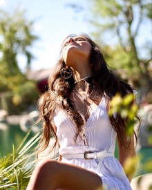 A woman with long hair gazes upward while sitting outdoors in a natural setting. She wears a white dress with lace details and a belt, accessorized with necklaces and large hoop earrings. The background features blurred green foliage and a body of water, giving a serene and summery feel.