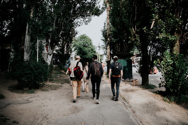 A group of people is walking along a tree-lined street. The individuals carry backpacks and appear to be casually dressed, suggesting a relaxed outing. The path is flanked by tall trees, providing a natural canopy. In the background, there is a small structure and a glimpse of a red car, indicating an urban setting.