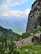 A scenic mountain trail with hikers walking along a gravel path. The landscape features lush greenery, towering rocky cliffs, and expansive views of distant mountain ranges under a partly cloudy blue sky.