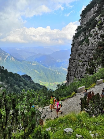 A scenic mountain trail with hikers walking along a gravel path. The landscape features lush greenery, towering rocky cliffs, and expansive views of distant mountain ranges under a partly cloudy blue sky.