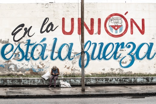 A person sits on a bench against a weathered wall with a large mural. The mural features bold letters in Spanish, partially obscured by a pole, and includes a round emblem with a star. The person's head is down, surrounded by bags, with a street and curb in the foreground.