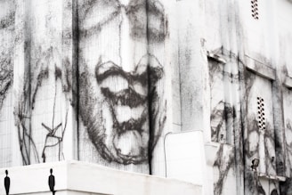 A large building façade featuring abstract and expressive black and white artwork. The artwork appears to depict a face with exaggerated features in an emotive style, adding a dramatic and thought-provoking element to the urban environment.