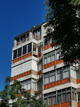 A multi-story residential building featuring a modernist design with white and orange accents. Large windows dominate the structure, providing a sense of openness. Lush green trees partially obscure the building, and the sky is clear and vivid blue.