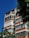 A multi-story residential building featuring a modernist design with white and orange accents. Large windows dominate the structure, providing a sense of openness. Lush green trees partially obscure the building, and the sky is clear and vivid blue.