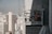 A construction worker in safety gear, including a helmet and orange vest, is positioned inside a suspended scaffold or work platform attached to the side of a tall building. The worker appears to be engaged in some maintenance or construction work. The background reveals a cityscape with numerous tall buildings and skyscrapers under a clear sky.