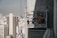 A construction worker in safety gear, including a helmet and orange vest, is positioned inside a suspended scaffold or work platform attached to the side of a tall building. The worker appears to be engaged in some maintenance or construction work. The background reveals a cityscape with numerous tall buildings and skyscrapers under a clear sky.
