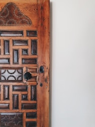 A close-up view of a wooden door featuring intricate carvings and geometric patterns. The door displays a combination of dark and light wood tones, with a noticeable aged texture. The right side of the image shows a plain white wall, adding contrast to the detailed woodwork.