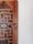 A close-up view of a wooden door featuring intricate carvings and geometric patterns. The door displays a combination of dark and light wood tones, with a noticeable aged texture. The right side of the image shows a plain white wall, adding contrast to the detailed woodwork.