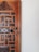 A close-up view of a wooden door featuring intricate carvings and geometric patterns. The door displays a combination of dark and light wood tones, with a noticeable aged texture. The right side of the image shows a plain white wall, adding contrast to the detailed woodwork.