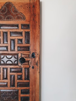 A close-up view of a wooden door featuring intricate carvings and geometric patterns. The door displays a combination of dark and light wood tones, with a noticeable aged texture. The right side of the image shows a plain white wall, adding contrast to the detailed woodwork.