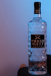 A large, transparent bottle with a textured, geometric design and a black label with gold lettering displaying the brand name 'Three Sixty Vodka'. The bottle is placed against a neutral-colored background, with a faint blue light casting a gentle hue over it.